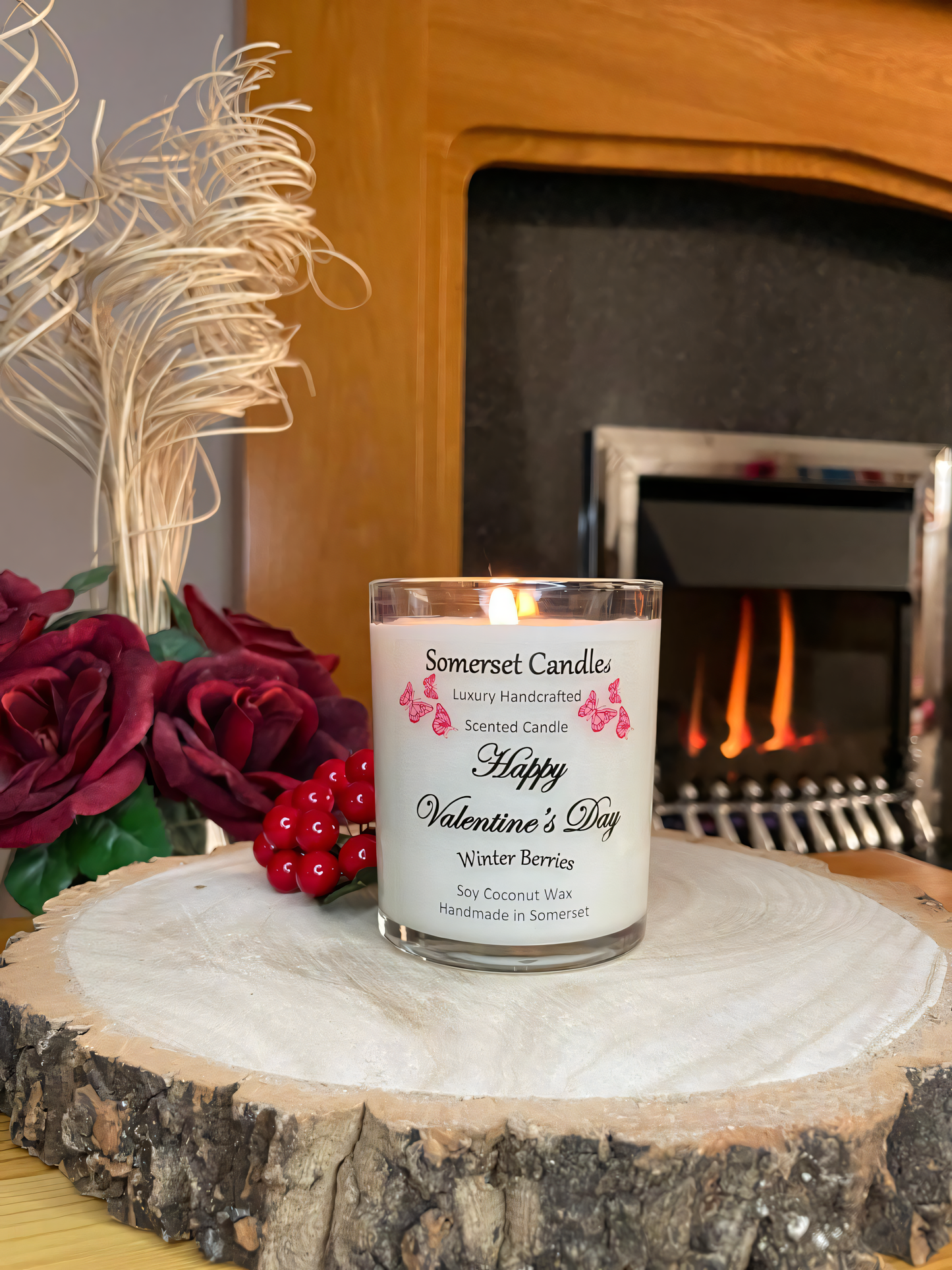 Valentines Day Candle Gift Winter Berries displayed on a wooden board with berries and roses. The candle is white in colour with bright red butterflies fluttering above the scent name. The candle is lit in front of a fire.