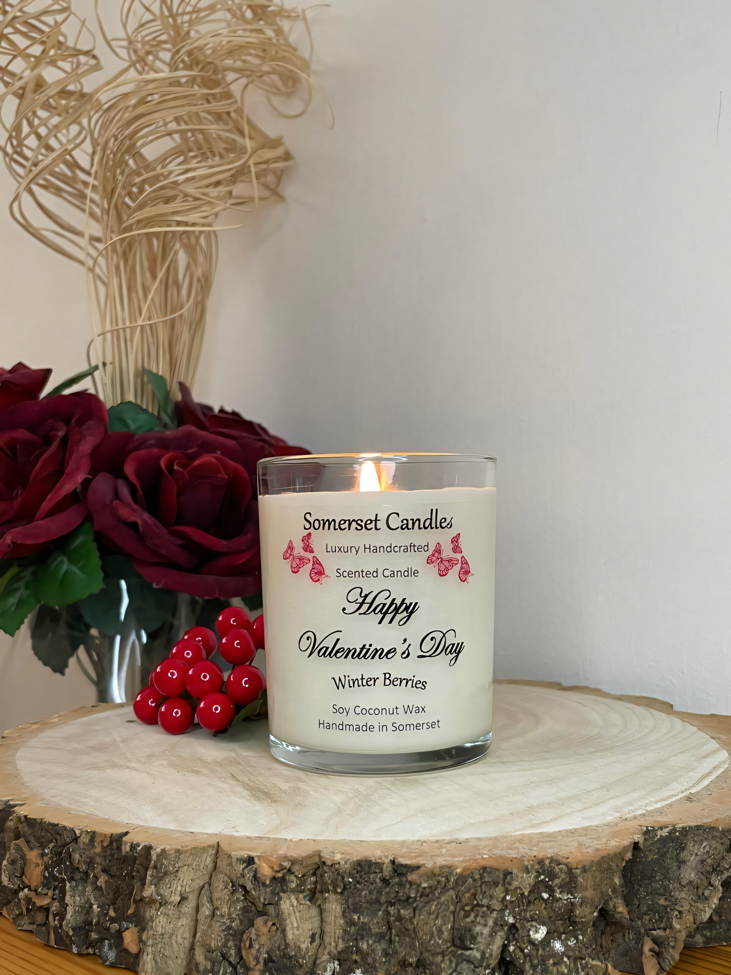 Valentines Day Candle Gift Winter Berries displayed on a wooden board with berries and roses. The candle is white in colour with bright red butterflies fluttering above the scent name. The candle is lit.