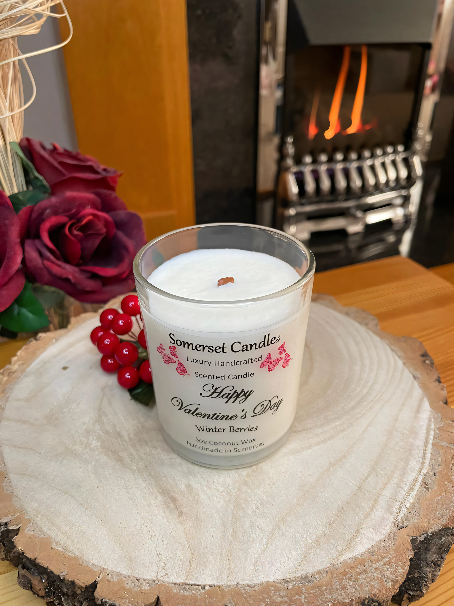 Valentines Day Candle Gift Winter Berries displayed on a wooden board with berries and roses. The candle is white in colour with bright red butterflies fluttering above the scent name. Viewed from above in front of the fireplace.