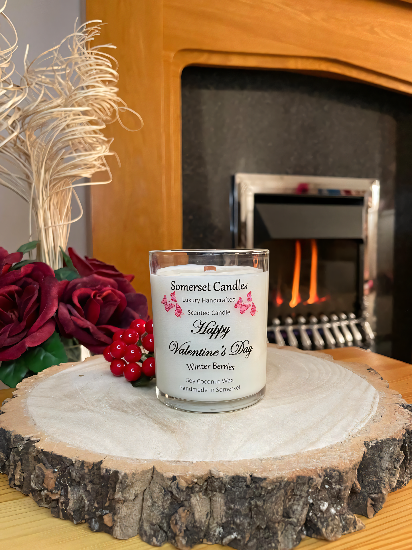 Valentines Day Candle Gift Winter Berries displayed on a wooden board with berries and roses. The candle is white in colour with bright red butterflies fluttering above the scent name. The candle is lit in front of a fire.