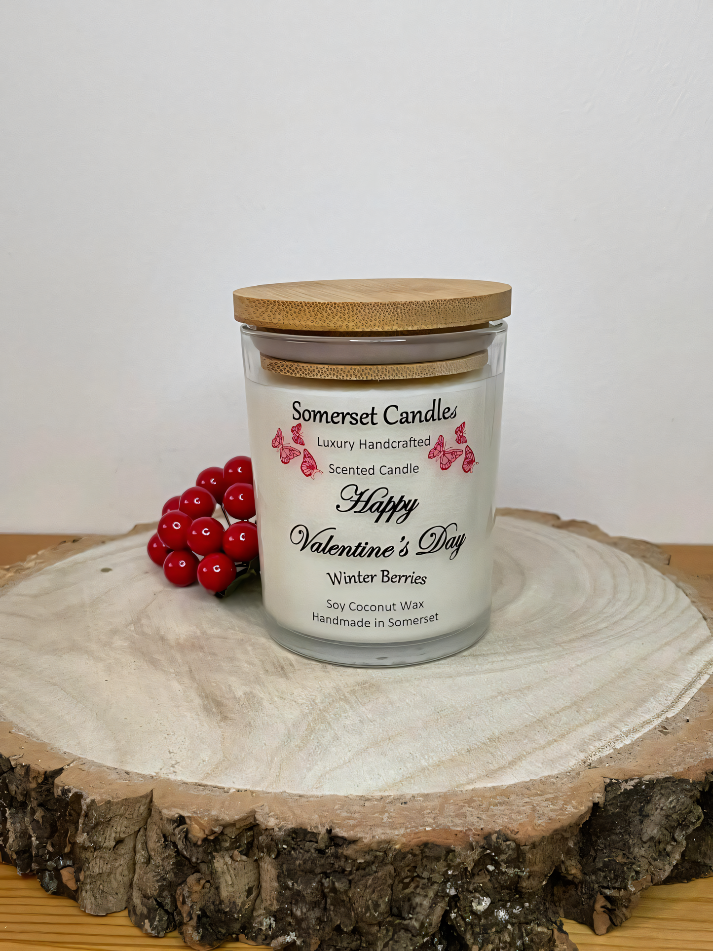 Valentines Day Candle Gift Winter Berries displayed on a wooden board with berries. The candle is white in colour with bright red butterflies fluttering above the scent name. A bamboo lid is fitted to the candle.
