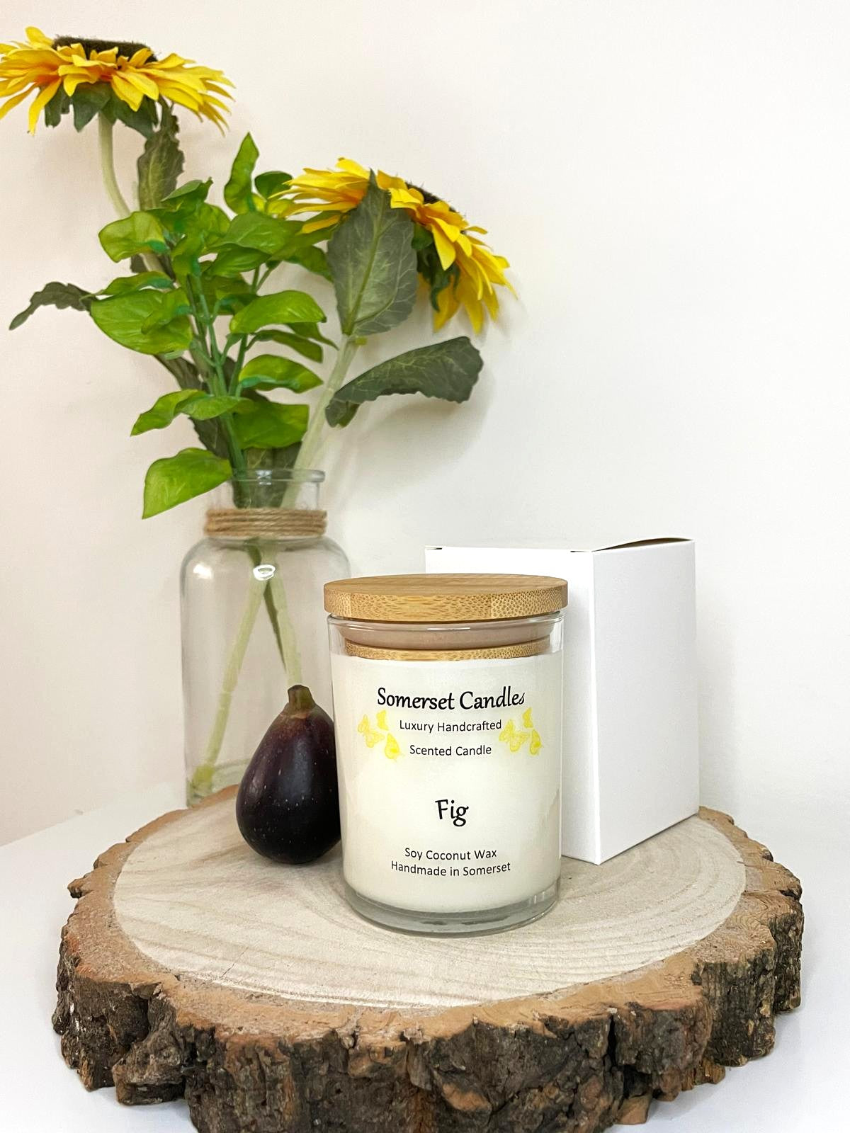 White soy candle in a 7oz clear glass jar with a natural wooden lid. The candle is made from a soy-coconut blend and scented with a fresh scent of ripe fig. Above the scent name on the label, several yellow butterflies are displayed, adding a romantic touch. The candle is presented with a plain white gift box. Clean, minimalist design ideal for gifts, home decor or self-care.