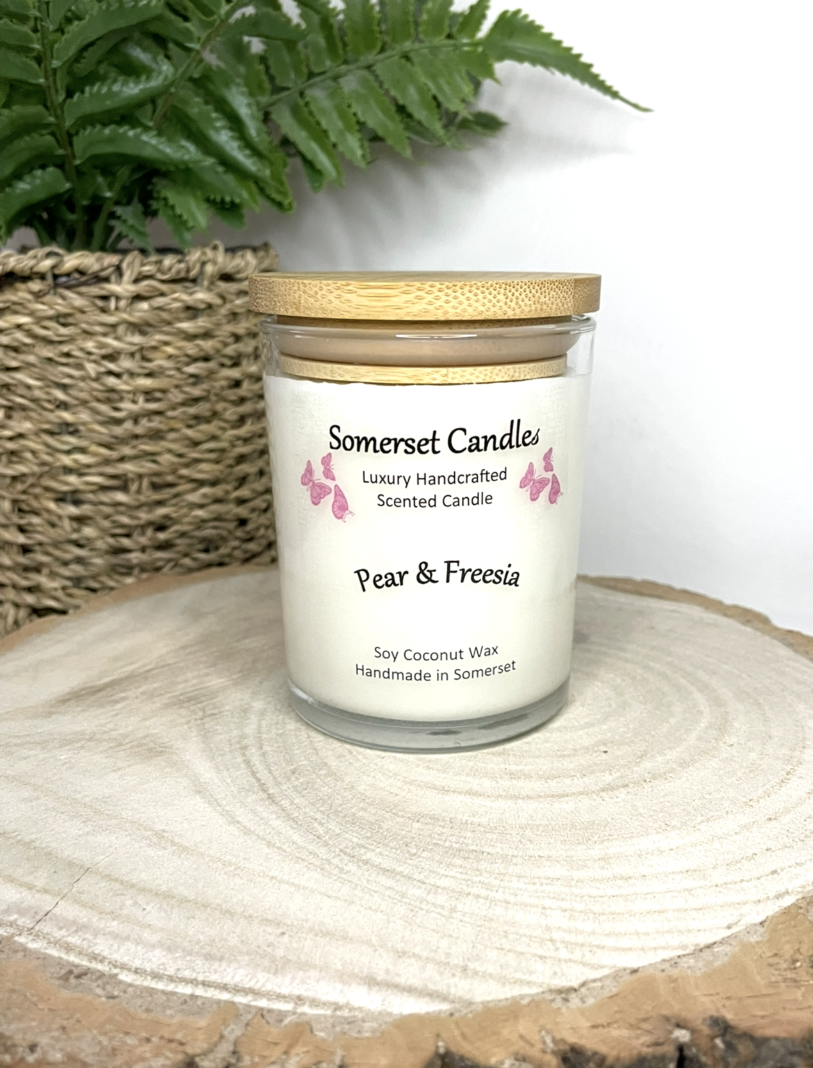 White soy candle in a 7oz clear glass jar with a natural wooden lid. The candle is made from a soy-coconut blend and scented with a delicate mix of pear and freesia. Above the scent name on the label, several pink butterflies are displayed, adding a romantic touch. Clean, minimalist design ideal for gifts, home decor or self-care.