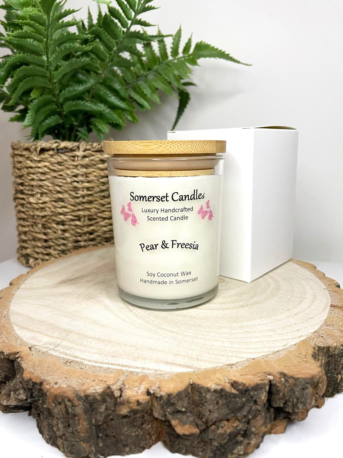 White soy candle in a 7oz clear glass jar with a natural wooden lid. The candle is made from a soy-coconut blend and scented with a delicate mix of pear and freesia. Above the scent name on the label, several pink butterflies are displayed, adding a romantic touch. The candle is presented with a plain white gift box. Clean, minimalist design ideal for gifts, home decor or self-care.