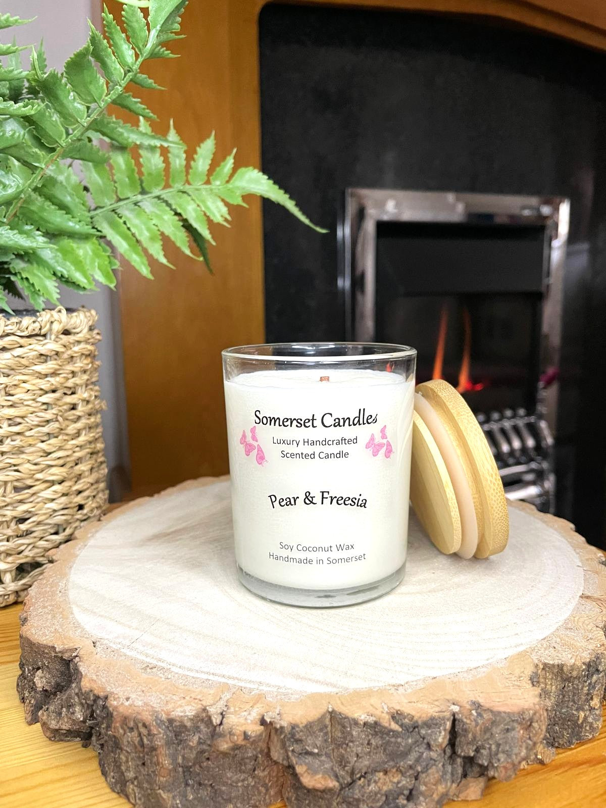 White soy candle in a 7oz clear glass jar with a natural wooden lid. The candle is made from a soy-coconut blend and scented with a delicate mix of pear and freesia. Above the scent name on the label, several pink butterflies are displayed, adding a romantic touch. The candle is in front of a lit fire. Clean, minimalist design ideal for gifts, home decor or self-care.
