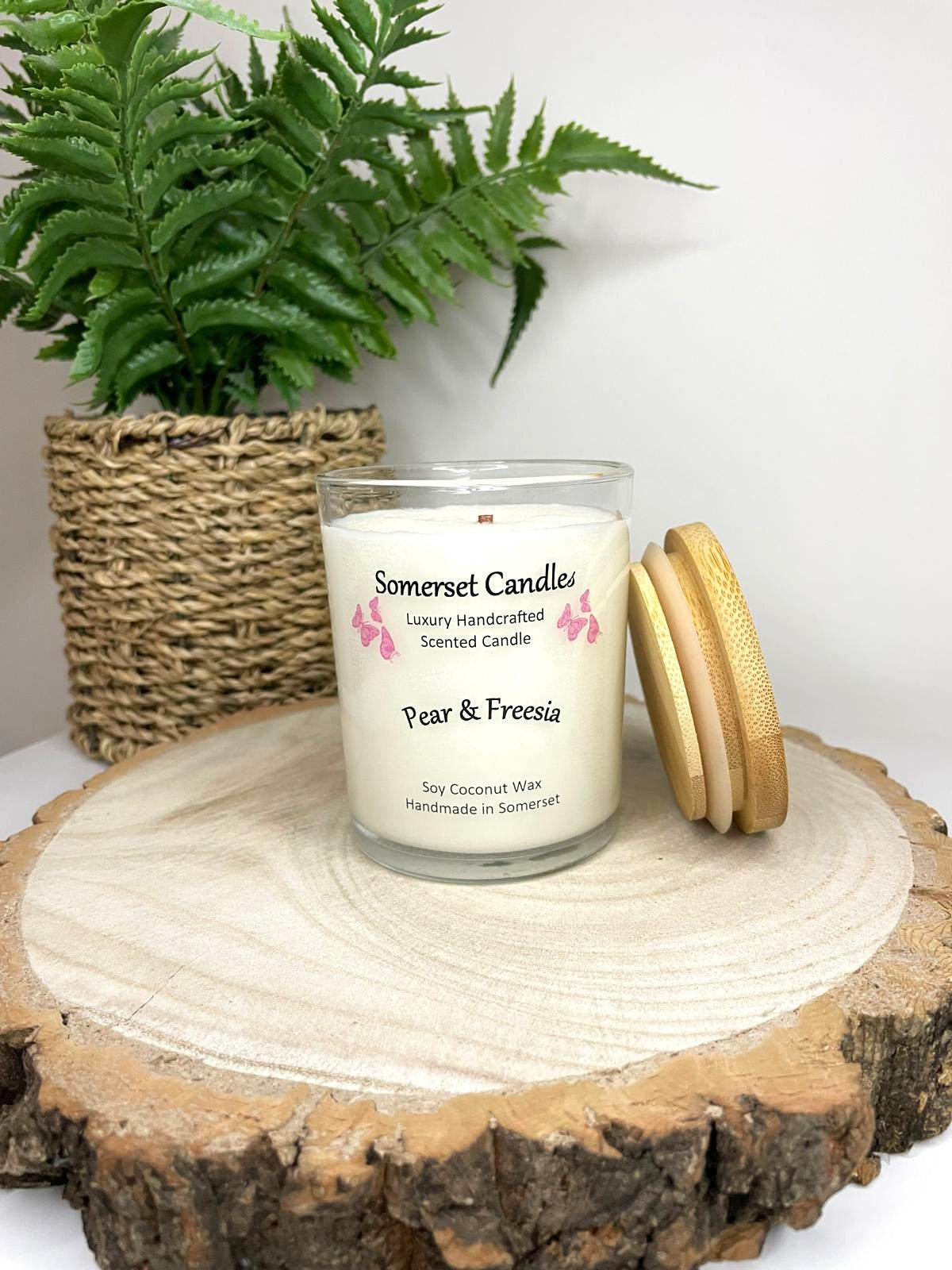 White soy candle in a 7oz clear glass jar with a natural wooden lid. The candle is made from a soy-coconut blend and scented with a delicate mix of pear and freesia. Above the scent name on the label, several pink butterflies are displayed, adding a romantic touch. Clean, minimalist design ideal for gifts, home decor or self-care.