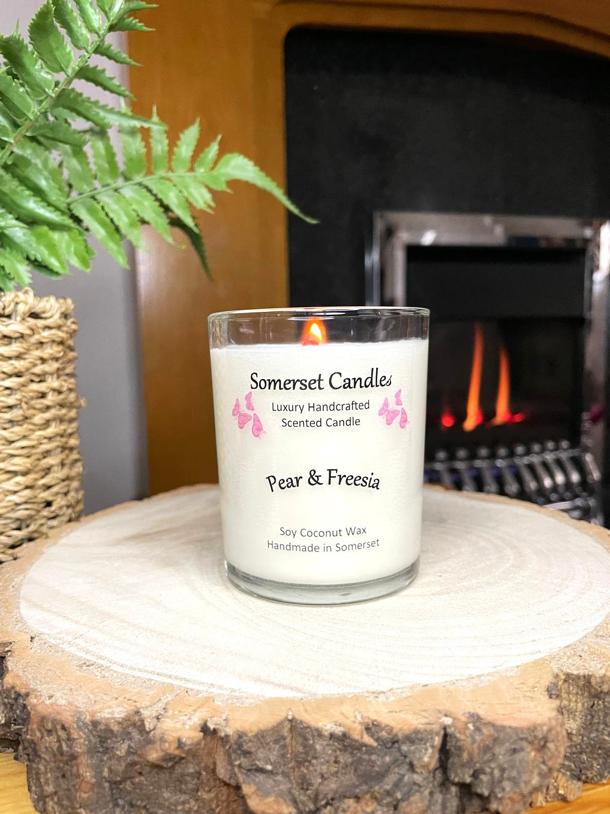 White soy candle in a 7oz clear glass jar with a natural wooden lid. The candle is made from a soy-coconut blend and scented with a delicate mix of pear and freesia. Above the scent name on the label, several pink butterflies are displayed, adding a romantic touch. The candle is lit in front of a lit fire place. Clean, minimalist design ideal for gifts, home decor or self-care.