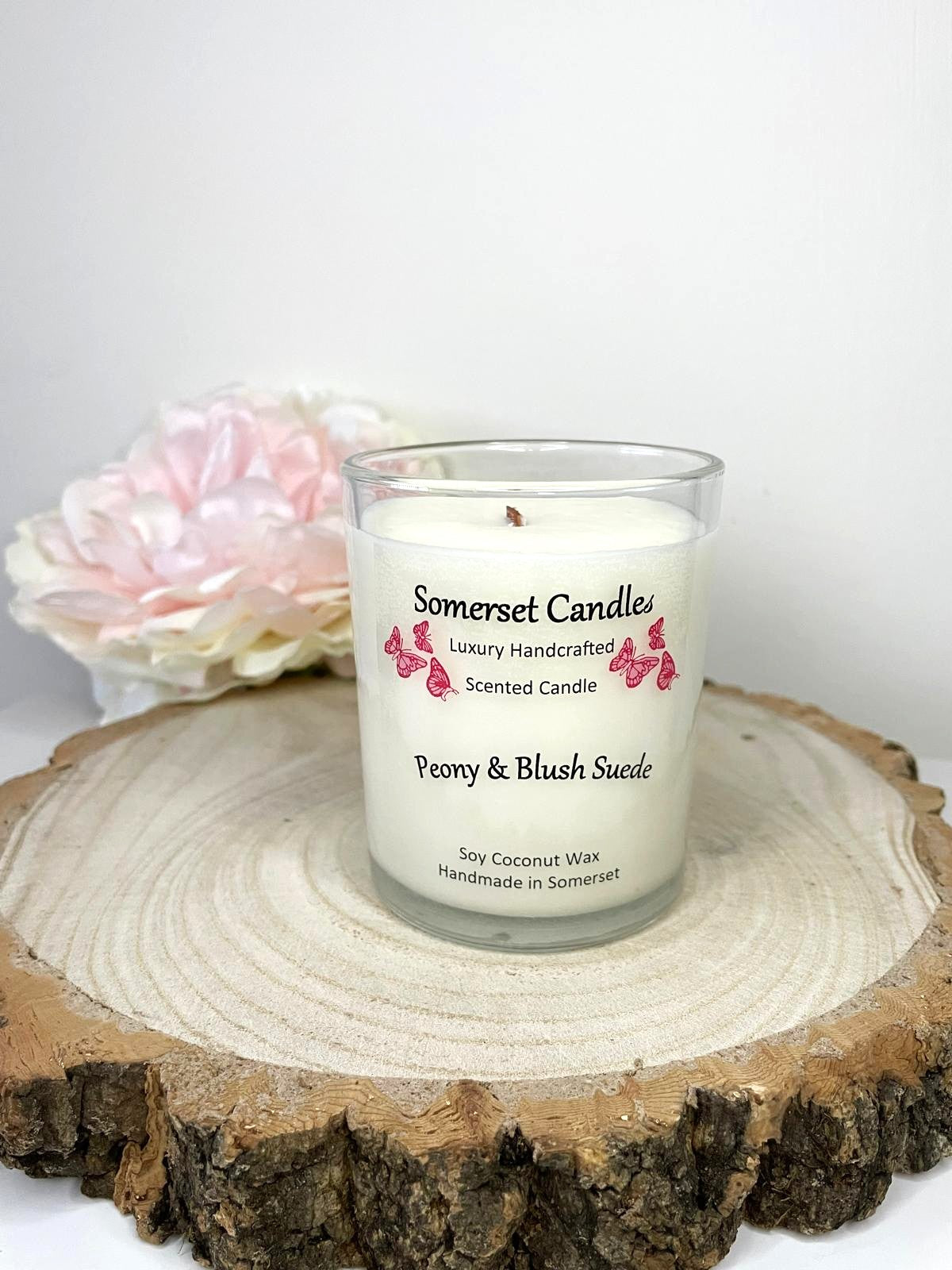 White soy candle in a 7oz clear glass jar with a natural wooden lid. The candle is made from a soy-coconut blend and scented with a soft elegant mix of Peony and Blush Suede. Above the scent name on the label, several pink butterflies are displayed, adding a romantic touch. Clean, minimalist design ideal for gifts, home decor or self-care.