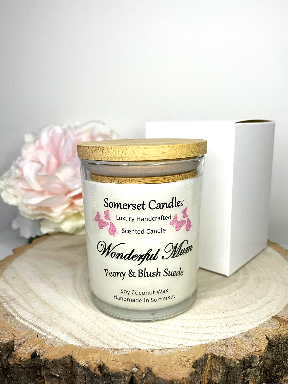 White soy candle in a 7oz clear glass jar with a natural wooden lid. The candle is made from a soy-coconut blend and scented with a delicate mix of peony and blush suede. Above the scent name on the label, several pink butterflies are displayed, adding a romantic touch. Clean, minimalist design ideal for gifts, home decor or self-care.