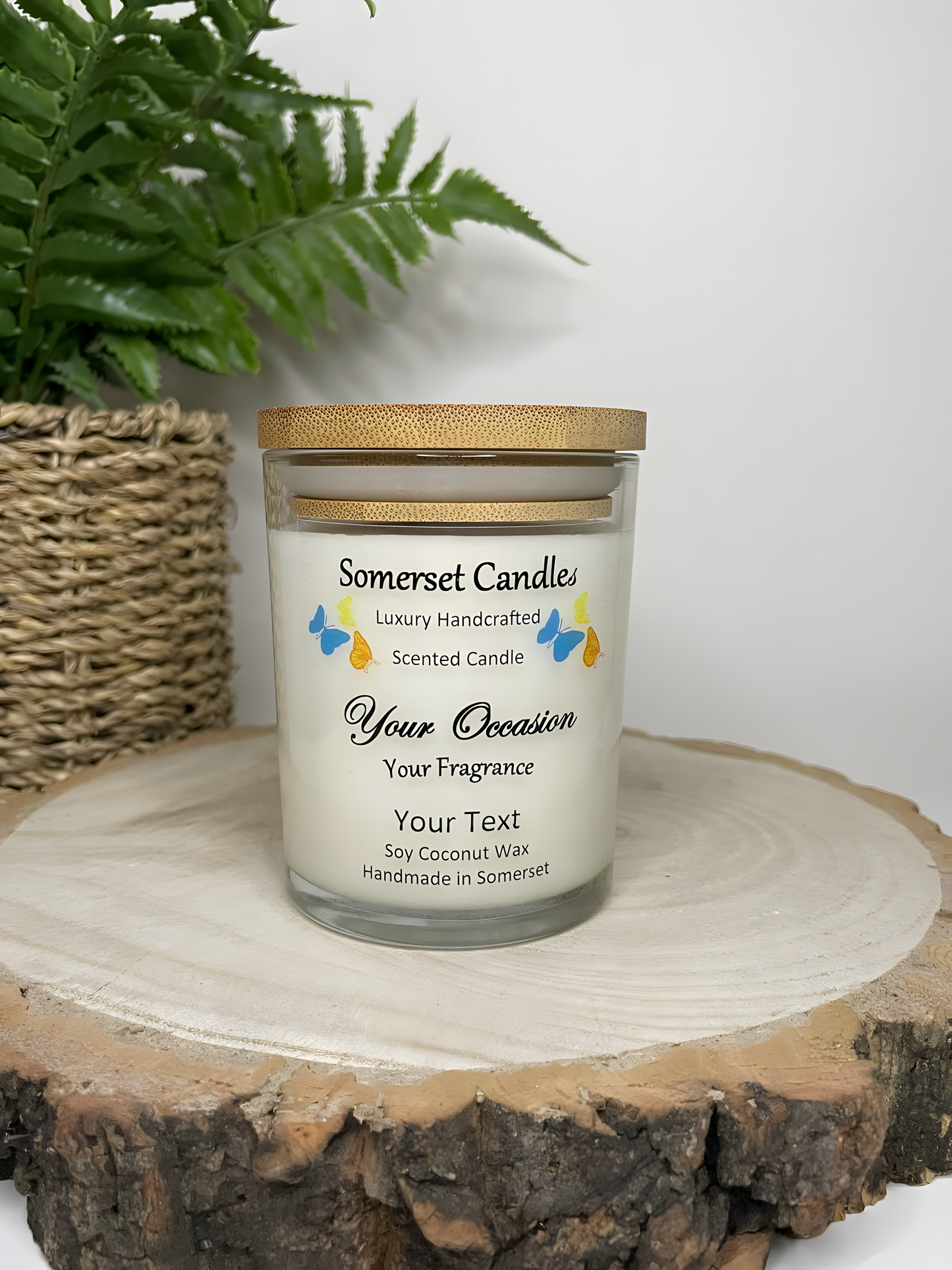 Personalised Scented Soy Candle displayed on a wooden log with a plant behind. The candle is white in colour with blue, orange & yellow butterflies above the scent name and a lovely bamboo lid.