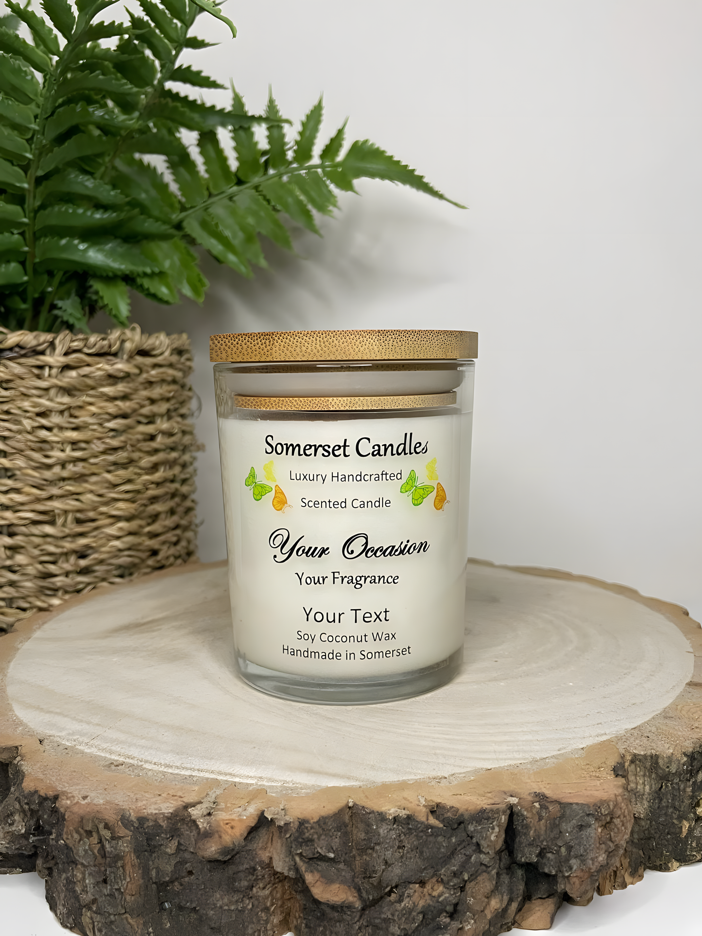 Personalised Scented Soy Candle displayed on a wooden log with a plant behind. The candle is white in colour with green, orange & yellow butterflies above the scent name and a lovely bamboo lid.
