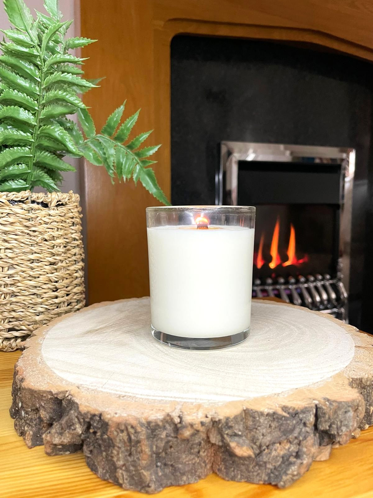White soy candle in a 7oz clear glass jar with a natural wooden lid. The candle is made from a soy-coconut blend and a variety of fresh fragrances. The candle has no label. The candle is lit in front of a lit fire. Clean, minimalist design ideal for gifts, home decor or self-care.