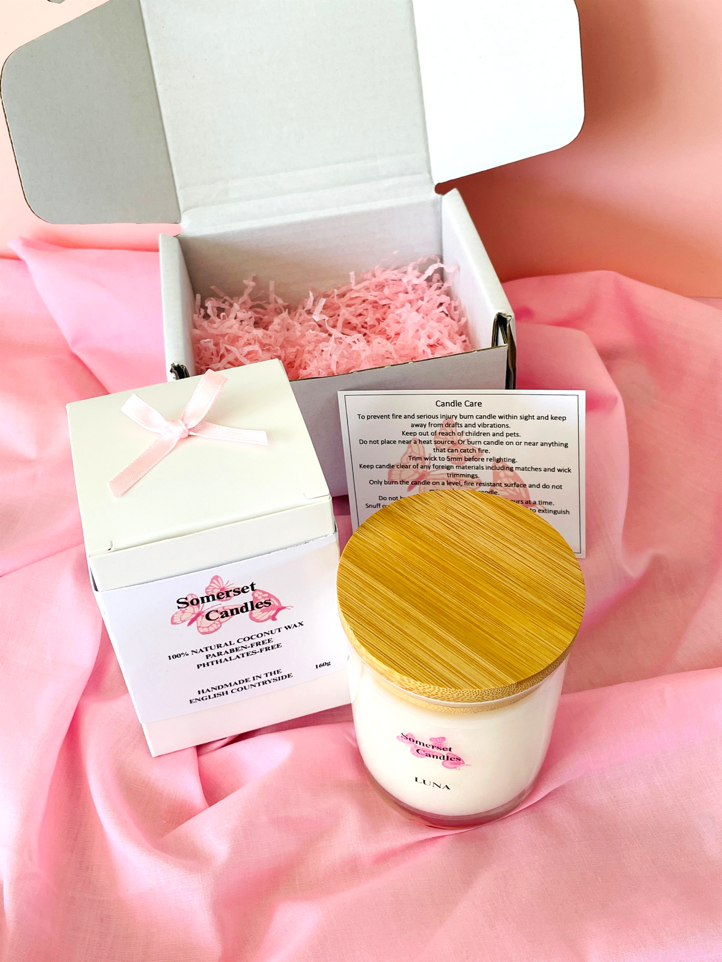 White candle with wooden lid in an open box on a pink fabric background
