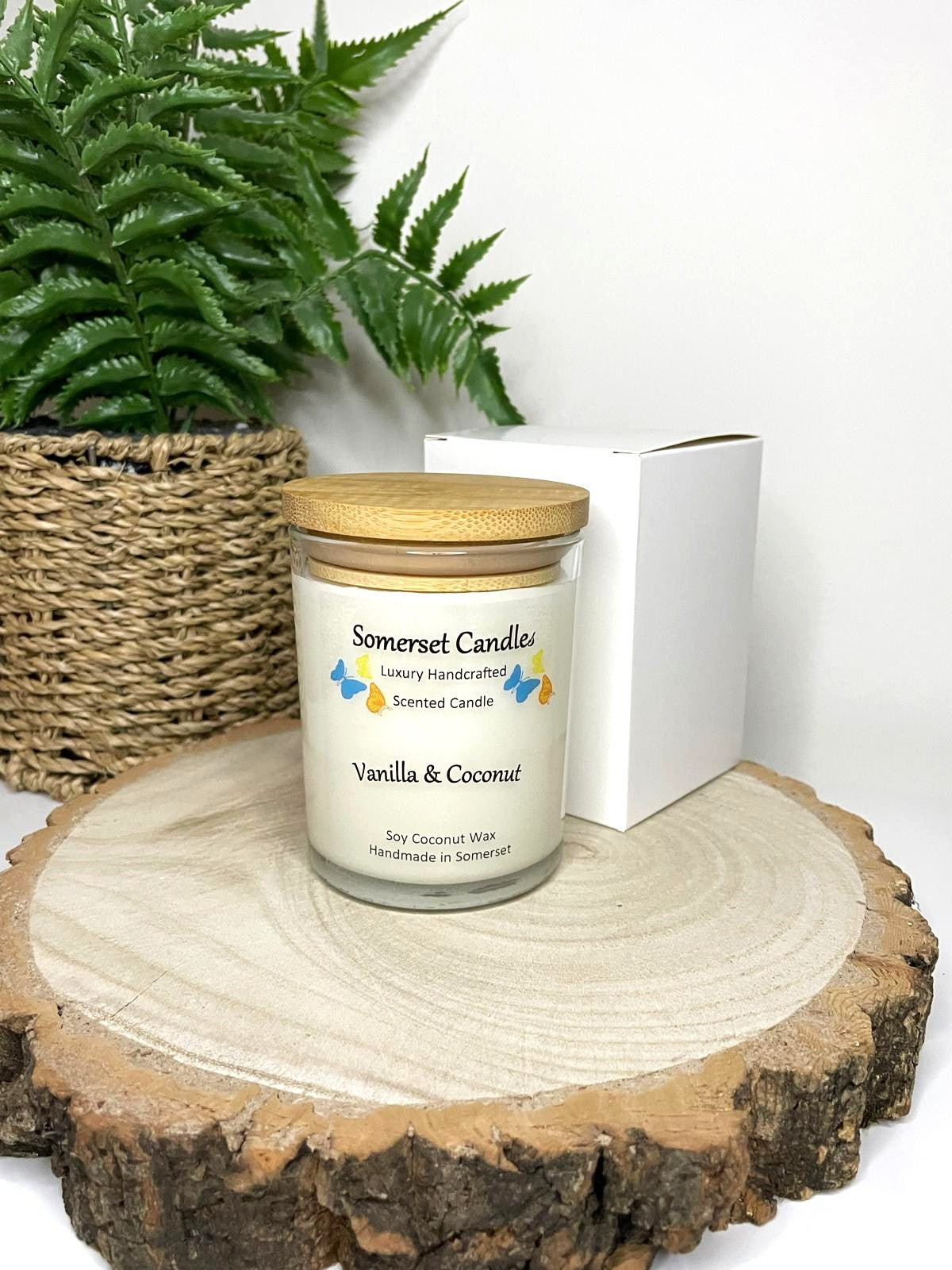 White soy candle in a 7oz clear glass jar with a natural wooden lid. The candle is made from a soy-coconut blend and scented with a fresh scent of vanilla and coconut. Above the scent name on the label, several blue, yellow and orange butterflies are displayed, adding a romantic touch. The candle is presented with a plain white gift box. Clean, minimalist design ideal for gifts, home decor or self-care.