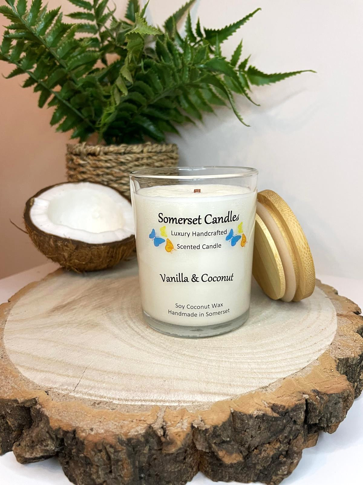 White soy candle in a 7oz clear glass jar with a natural wooden lid. The candle is made from a soy-coconut blend and scented with a fresh scent of vanilla and coconut. Above the scent name on the label, several blue, yellow and orange butterflies are displayed, adding a romantic touch. Clean, minimalist design ideal for gifts, home decor or self-care.