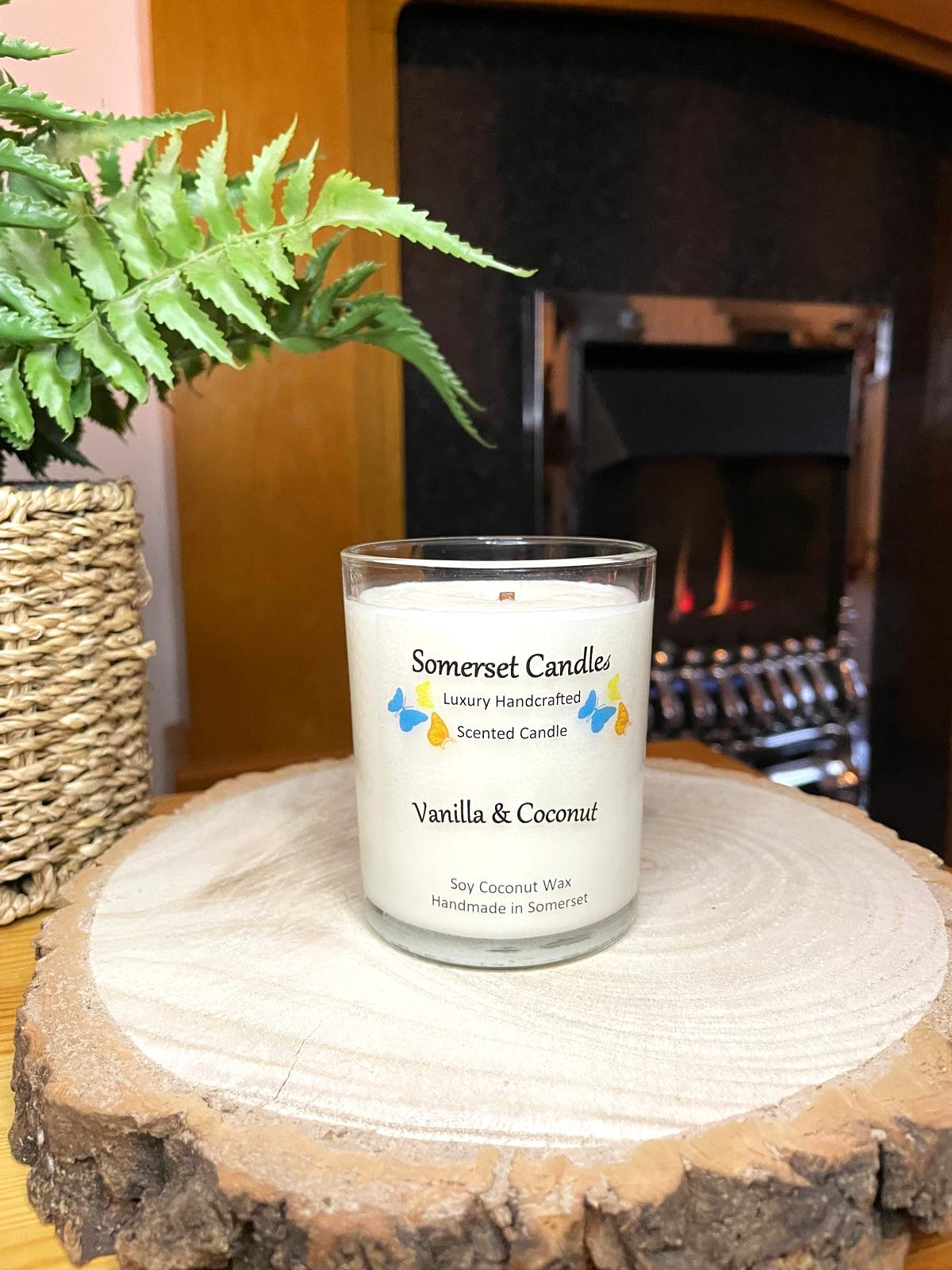 White soy candle in a 7oz clear glass jar with a natural wooden lid. The candle is made from a soy-coconut blend and scented with a fresh scent of vanilla and coconut. Above the scent name on the label, several blue, yellow and orange butterflies are displayed, adding a romantic touch. Clean, minimalist design ideal for gifts, home decor or self-care.