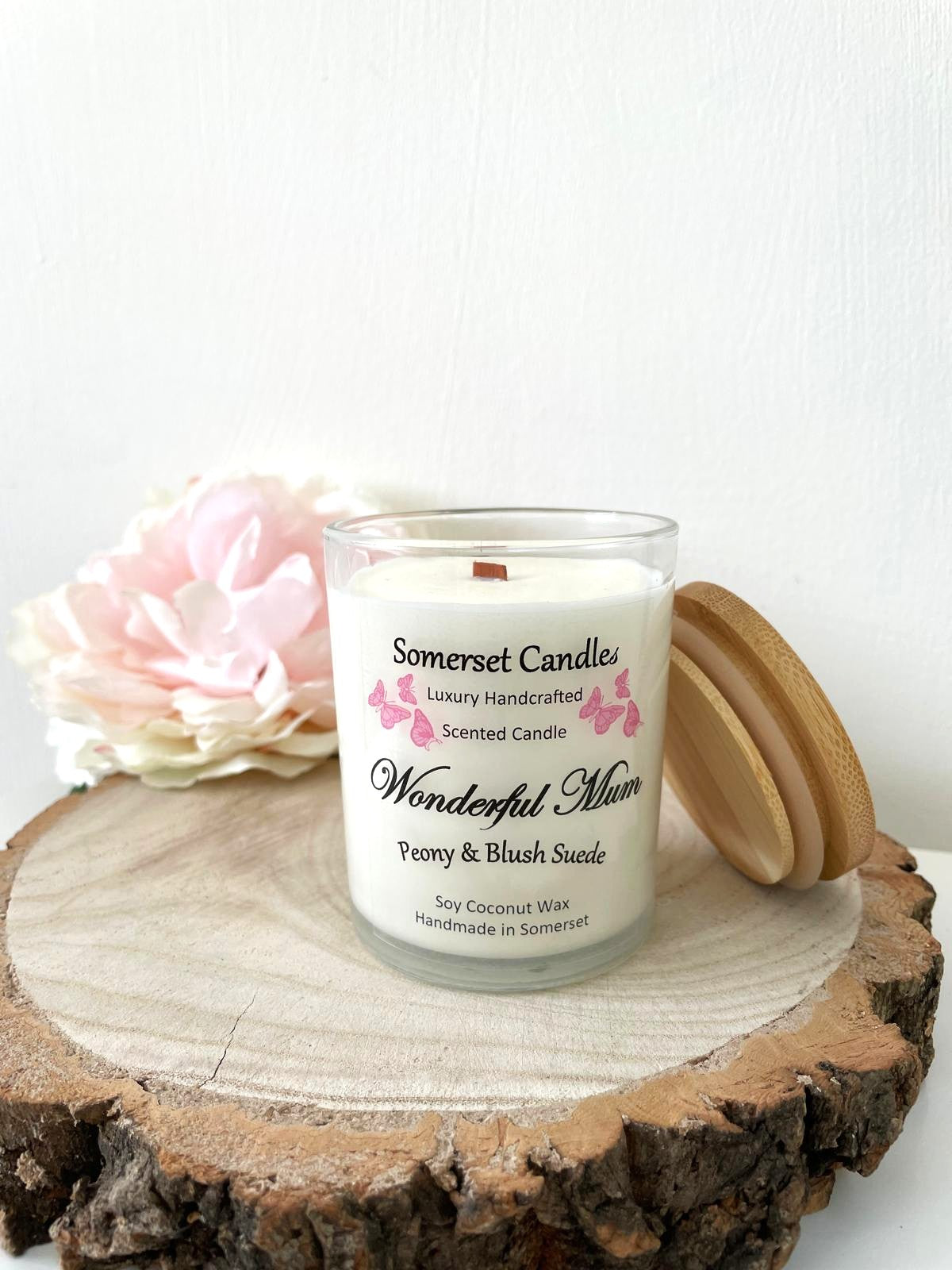 White soy candle in a 7oz clear glass jar with a natural wooden lid. The candle is made from a soy-coconut blend and scented with a delicate mix of peony and blush suede. Above the scent name on the label, several pink butterflies are displayed, adding a romantic touch. Clean, minimalist design ideal for gifts, home decor or self-care.