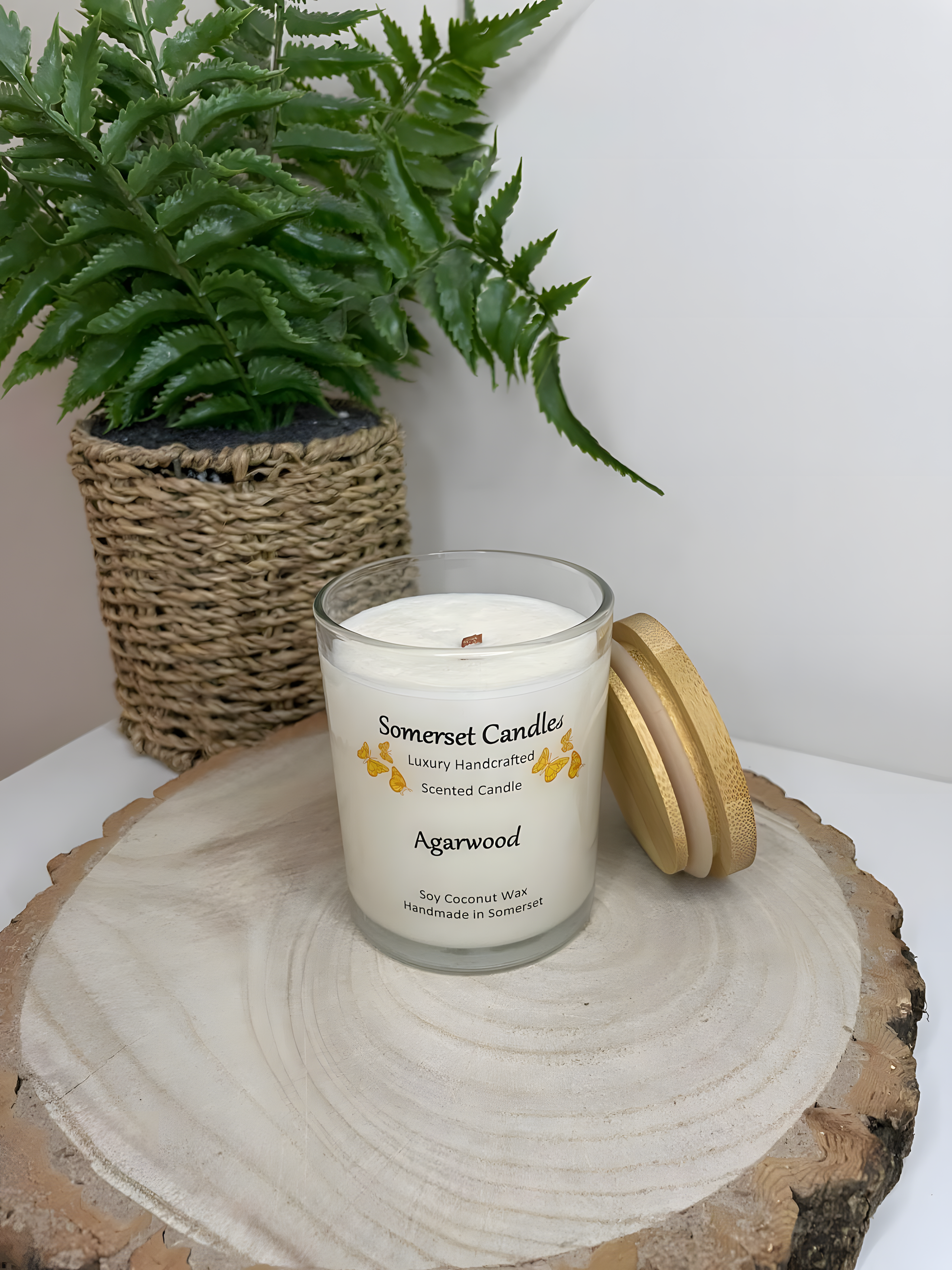 Agarwood soy candle displayed on a wooden board. The candle is white in colour with bright orange butterflies above the scent name. There is a lovely bamboo lid resting to the side of the clear glass candle jar. The non toxic crackling wooden wick can be seen.