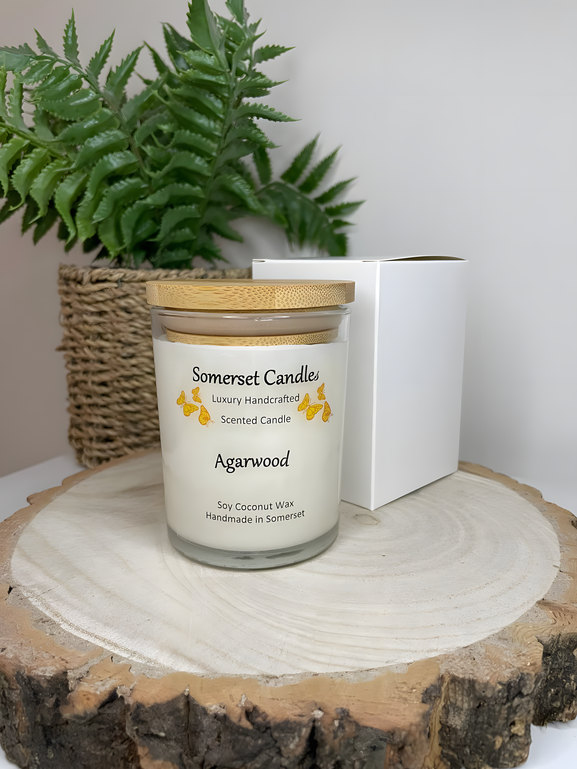 Agarwood soy candle displayed on a wooden board with a white gift box. The candle is white in colour with bright orange butterflies above the scent name with a lovely bamboo lid. The candle jar is clear glass.