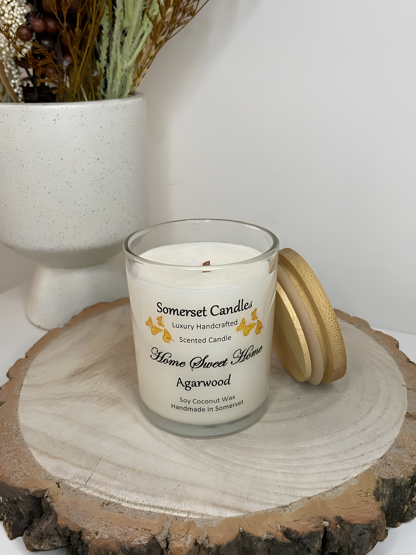 Home Sweet Home Agarwood soy candle is displayed on a wooden board with autumn flowers. The candle is white in colour with orange butterflies fluttering above the scent name and fitted with a lovely bamboo lid.