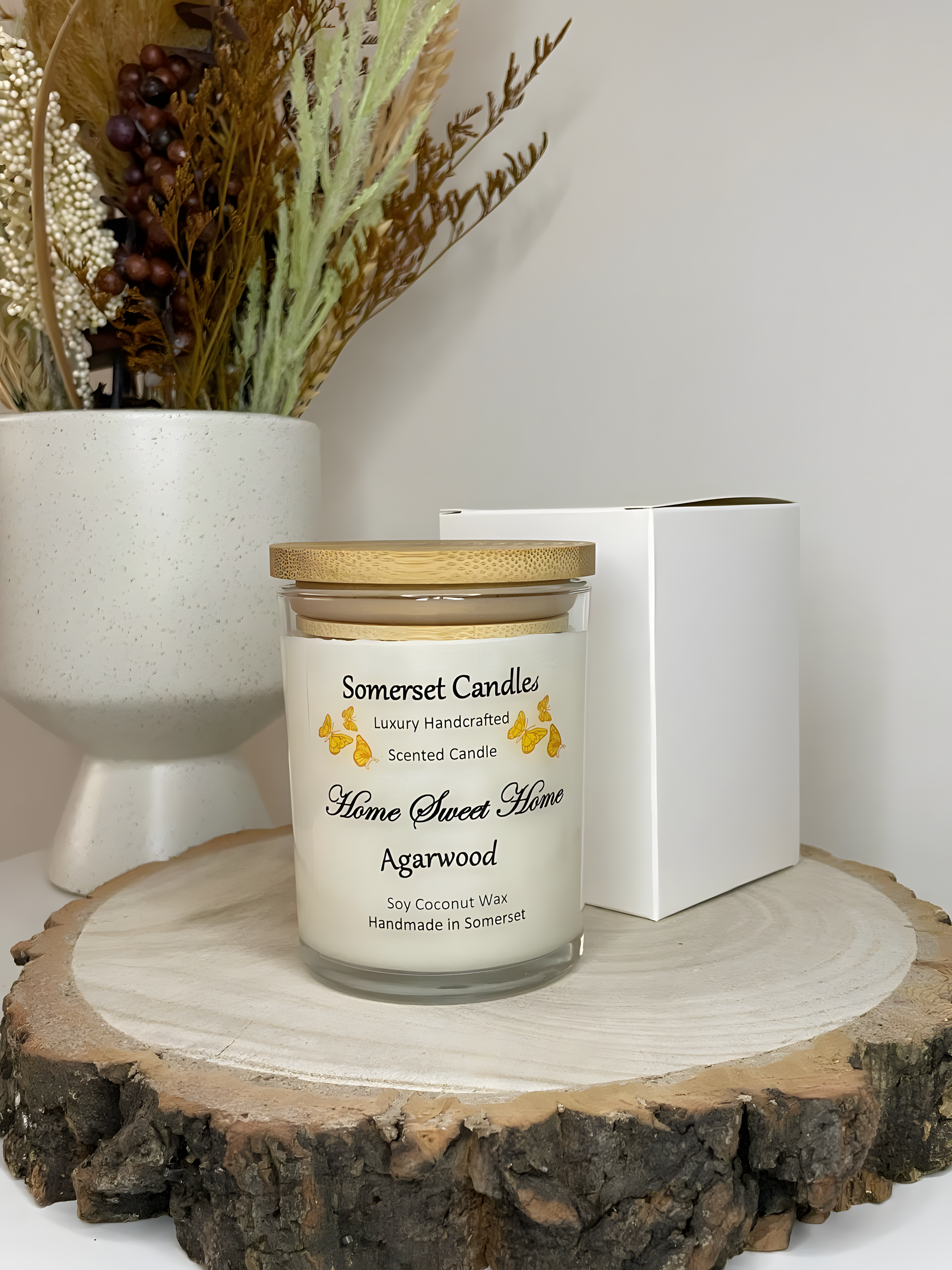 Home Sweet Home Agarwood soy candle is displayed on a wooden board with autumn flowers and a gift box. The candle is white in colour with orange butterflies fluttering above the scent name and fitted with a lovely bamboo lid.