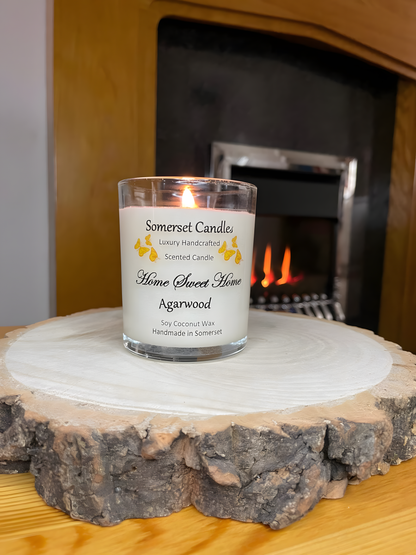 Home Sweet Home Agarwood soy candle is displayed on a wooden board with autumn flowers in front of a lit fire. The candle is white in colour with orange butterflies fluttering above the scent name.