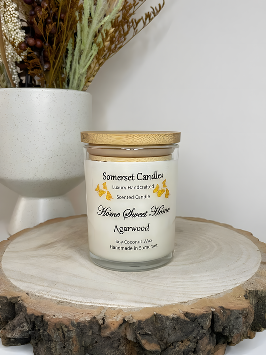 Home Sweet Home Agarwood soy candle is displayed on a wooden board with autumn flowers. The candle is white in colour with orange butterflies fluttering above the scent name and fitted with a lovely bamboo lid.