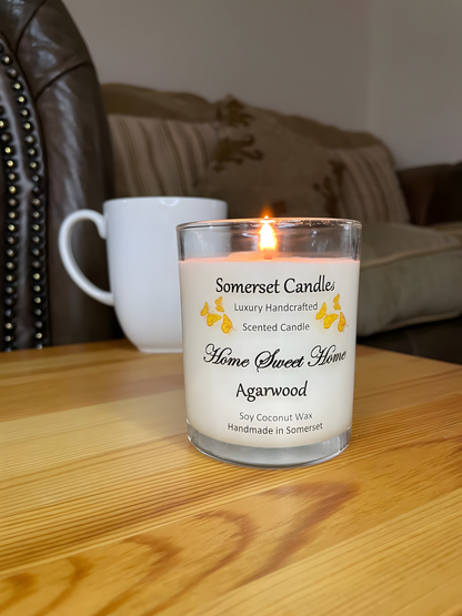 Home Sweet Home Agarwood soy candle is displayed on a wooden table with a mug. The candle is white in colour with orange butterflies fluttering above the scent name.