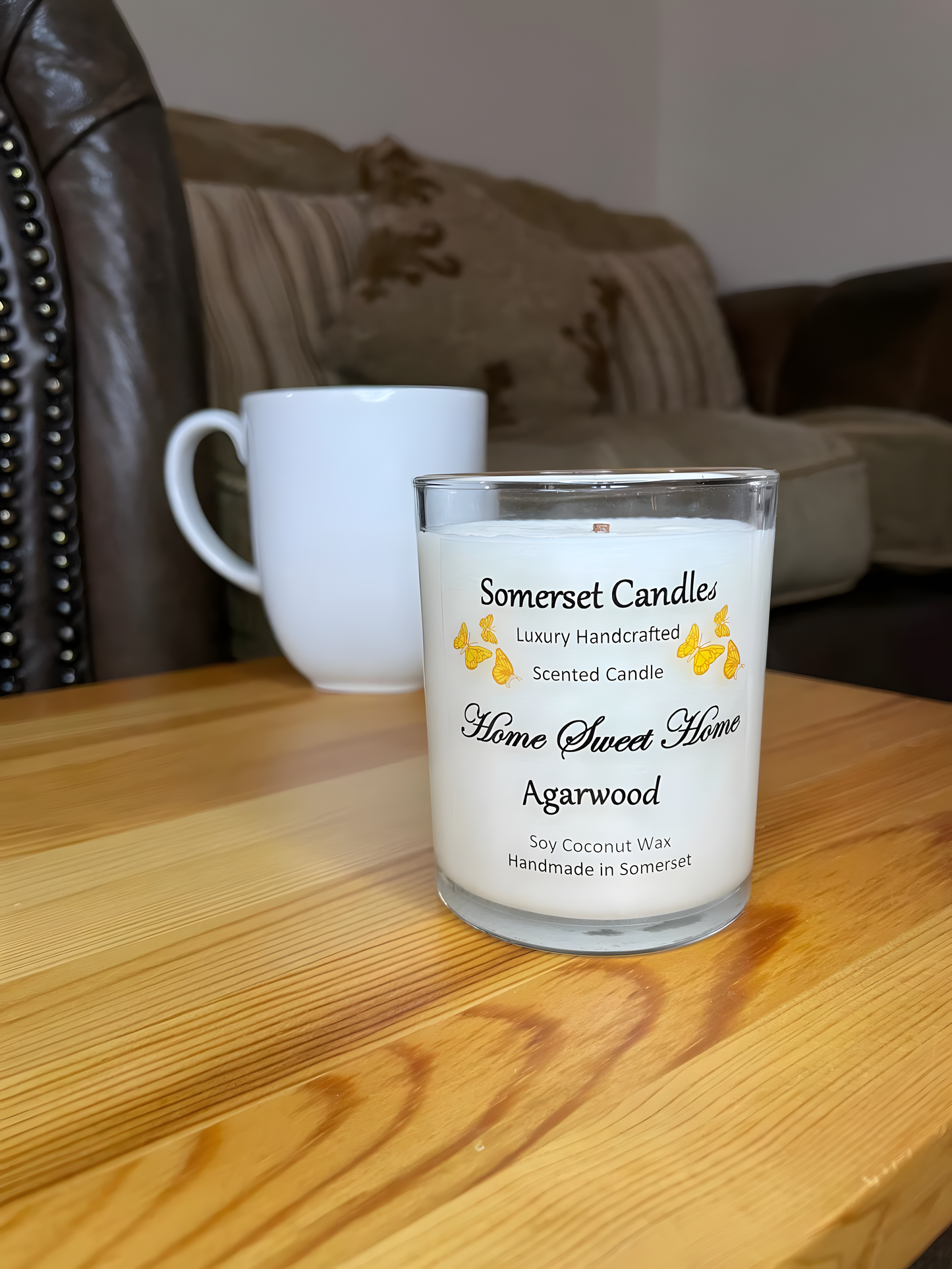 Home Sweet Home Agarwood soy candle is displayed on a wooden table with a mug. The candle is white in colour with orange butterflies fluttering above the scent name.