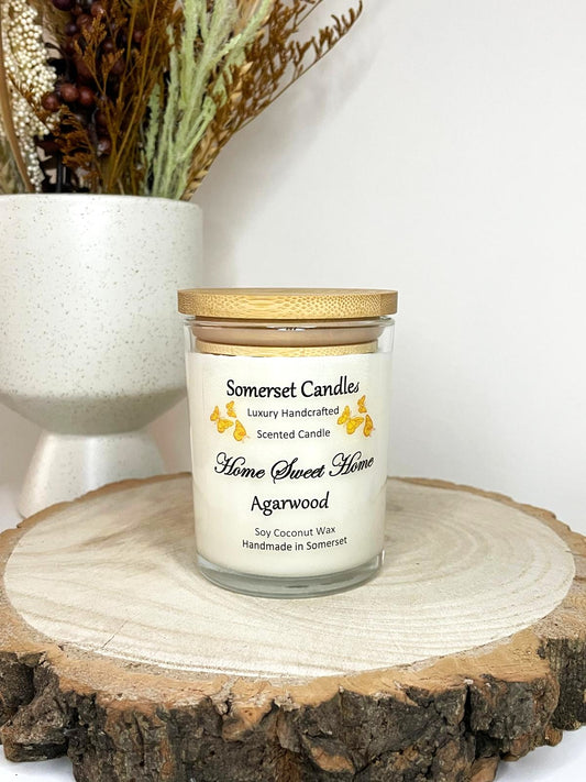 White soy candle in a 7oz clear glass jar with a natural wooden lid. The candle is made from a soy-coconut blend and scented with agarwod. Above the scent name on the label, several Orange butterflies are displayed, adding a romantic touch. Clean, minimalist design ideal for gifts, home decor or self-care.