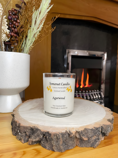 Agarwood soy candle displayed on a wooden board in front of a lit fire. The candle is white in colour with bright orange butterflies above the scent name. The non toxic crackling wooden wick can be seen and the candle jar is clear glass.