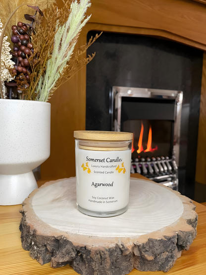 Agarwood soy candle displayed on a wooden board in front of a lit fire. The candle is white in colour with bright orange butterflies above the scent name with a lovely bamboo lid. The candle jar is clear glass.