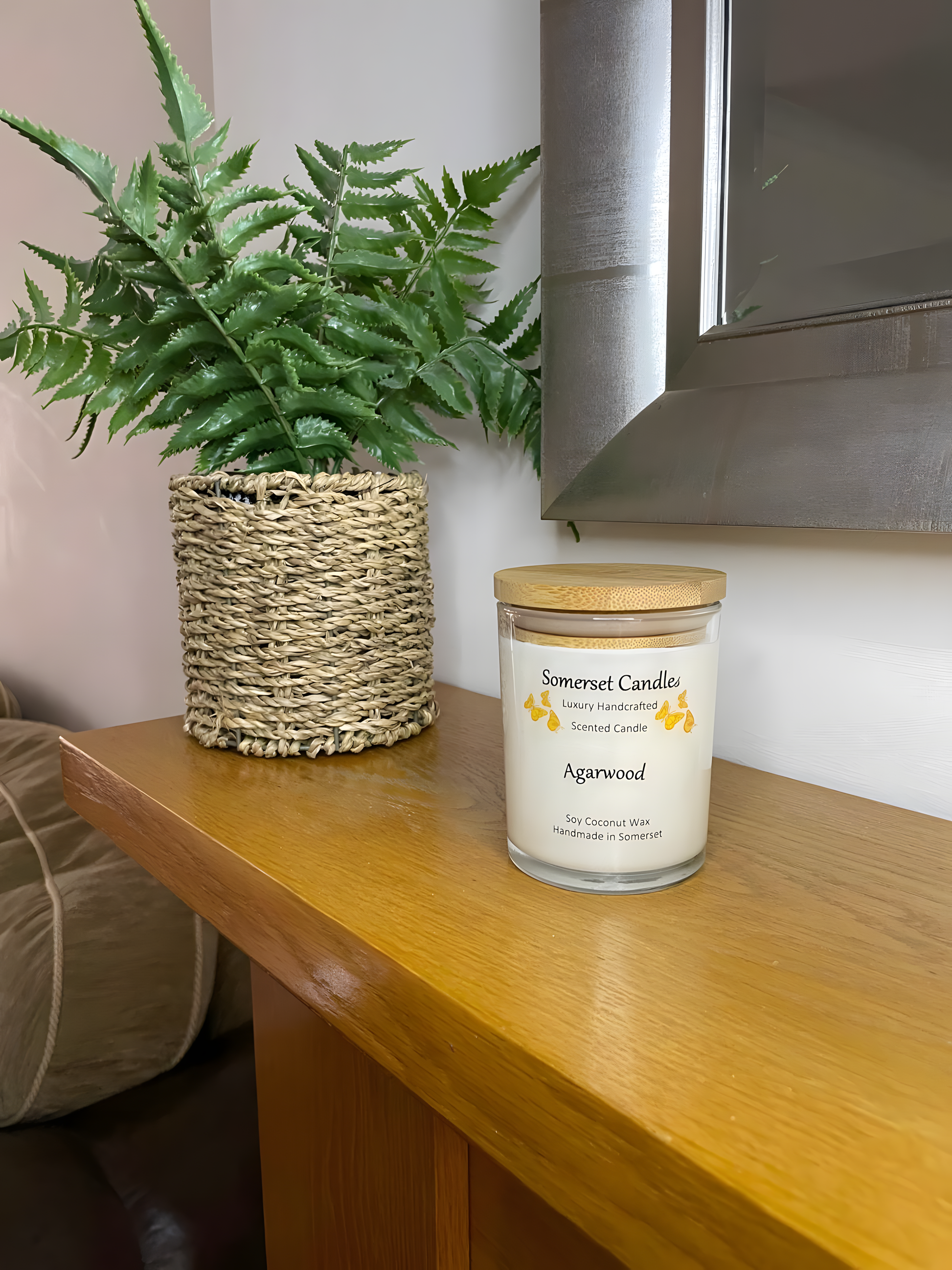 Agarwood soy candle displayed on a wooden mantel piece. The candle is white in colour with bright orange butterflies above the scent name with a lovely bamboo lid. The candle jar is clear glass.