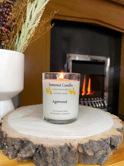 Agarwood soy candle displayed on a wooden board in front of a lit fire. The candle is white in colour with bright orange butterflies above the scent name. The candle is lit and has a clear glass container.