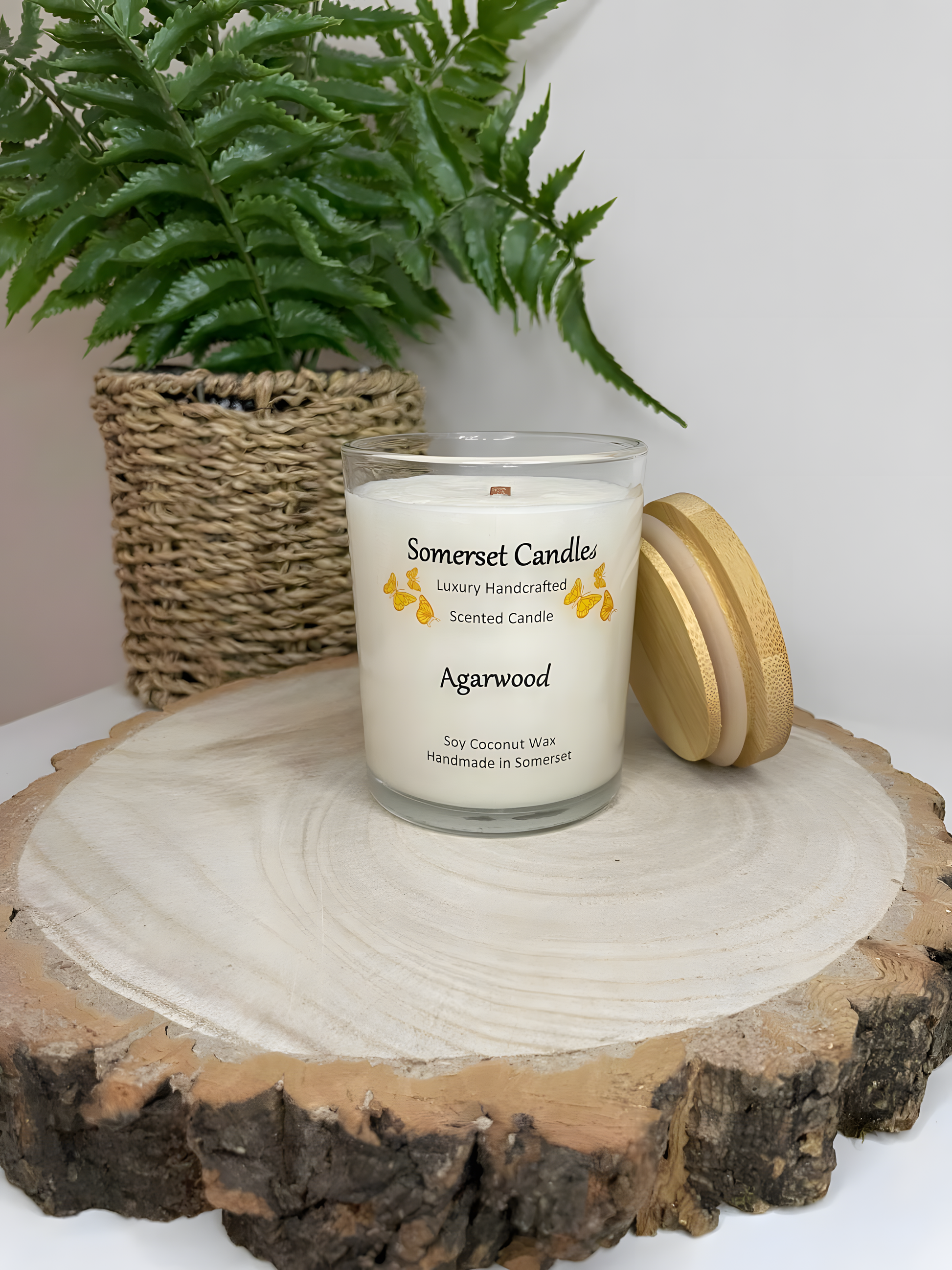 Agarwood soy candle displayed on a wooden board. The candle is white in colour with bright orange butterflies above the scent name. There is a lovely bamboo lid resting against the side of the glass candle container.