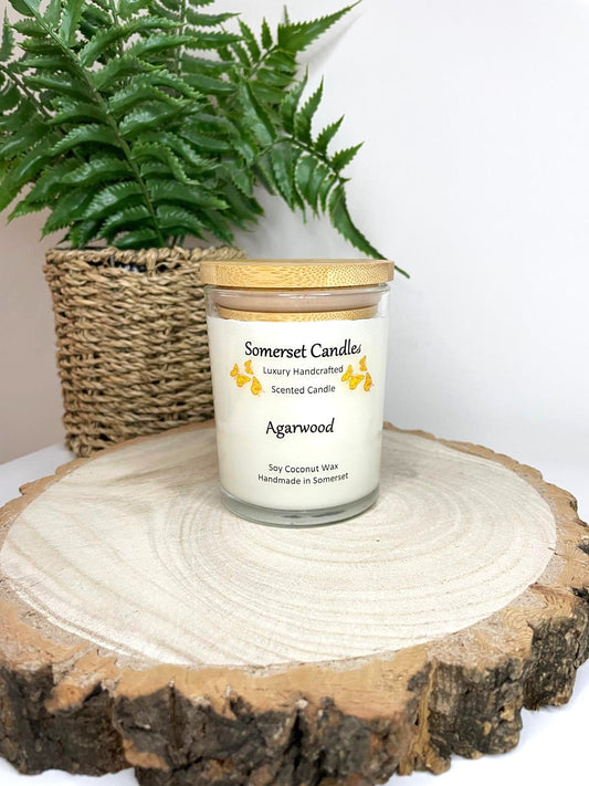 White soy candle in a 7oz clear glass jar with a natural wooden lid. The candle is made from a soy-coconut blend and scented with woodsie scent of agarwood. Above the scent name on the label, several orange butterflies are displayed, adding a romantic touch. Clean, minimalist design ideal for gifts, home decor or self-care.