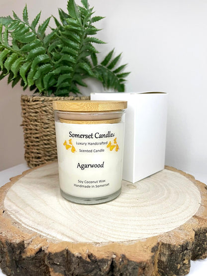 White soy candle in a 7oz clear glass jar with a natural wooden lid. The candle is made from a soy-coconut blend and scented with woodsie scent of agarwood. Above the scent name on the label, several orange butterflies are displayed, adding a romantic touch. The candle is presented with a plain white gift box. Clean, minimalist design ideal for gifts, home decor or self-care.
