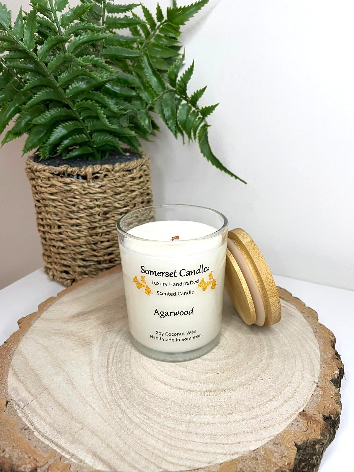 White soy candle in a 7oz clear glass jar with a natural wooden lid. The candle is made from a soy-coconut blend and scented with woodsie scent of agarwood. Above the scent name on the label, several orange butterflies are displayed, adding a romantic touch. Clean, minimalist design ideal for gifts, home decor or self-care.