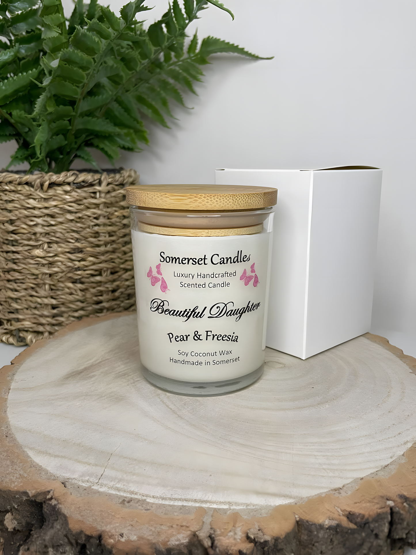 Beautiful Daughter pear & freesia scented soy candle   on a wooden board with a white gift box. The candle is white in colour with bright pink butterflies fluttering above the scent name and fitted with a lovely bamboo lid.