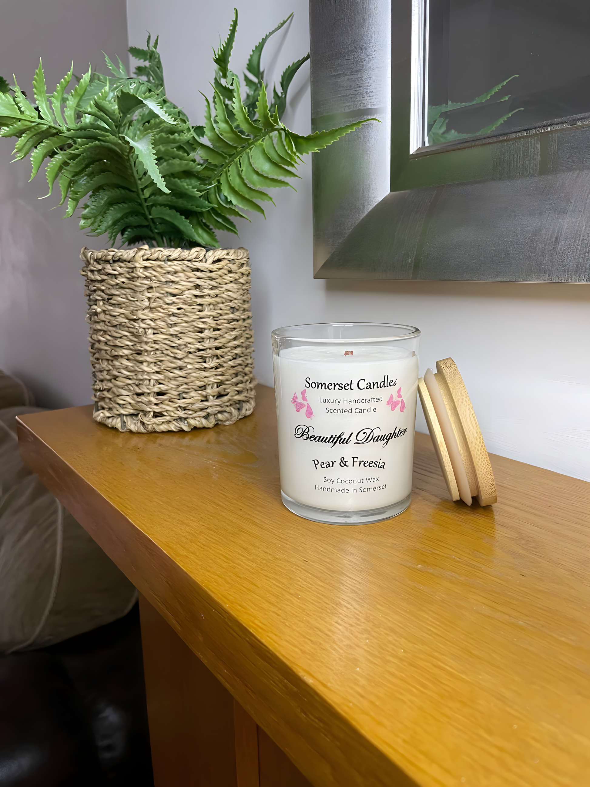 Beautiful Daughter Pear & Freesia scented soy candle  on the mantel piece. The candle is white in colour with bright pink butterflies fluttering above the scent name with a lovely bamboo lid. The candle jar is clear glass.