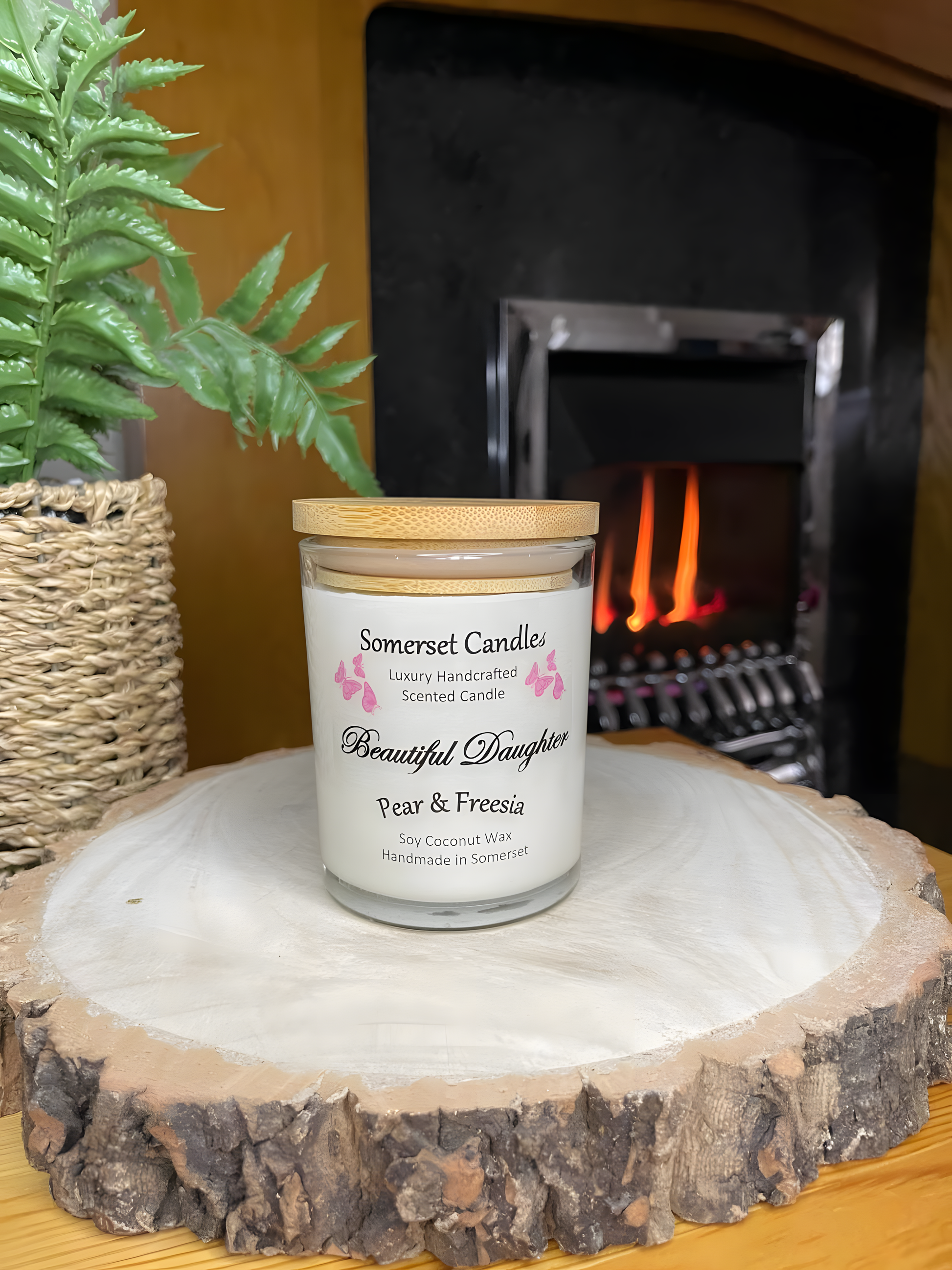 Beautiful Daughter Pear & Freesia scented soy candle  on a wooden board in front of a lit fire. The candle is white in colour with bright pink butterflies fluttering above the scent name and fitted with a lovely bamboo lid.