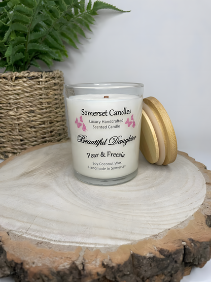 Beautiful Daughter pear & freesia  scented soy candle is displayed on a wooden board. The candle is white in colour with bright pink butterflies fluttering above the scent name with a lovely bamboo lid resting against the 
clear glass candle jar.