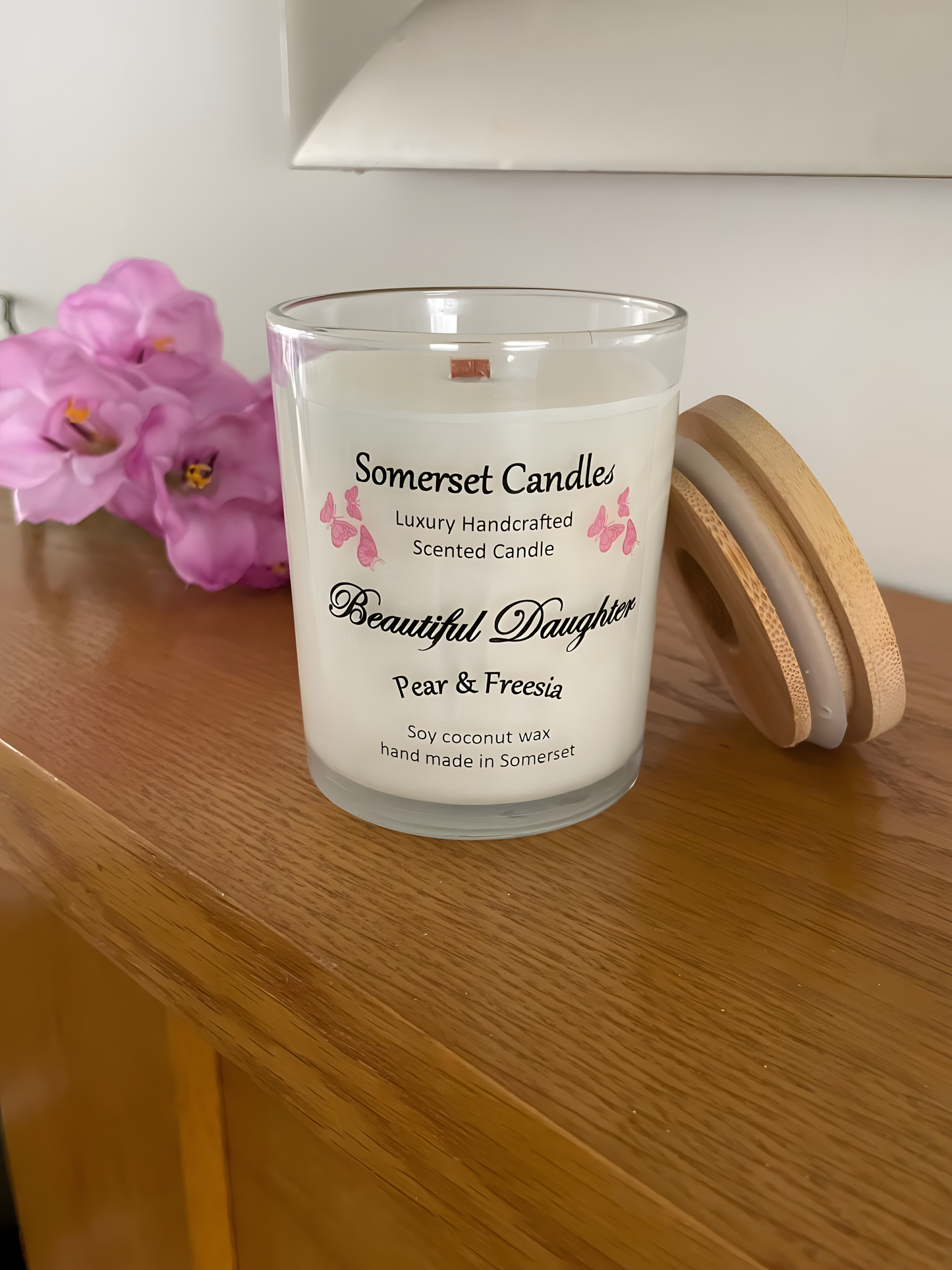 Beautiful Daughter pear & freesia scented soy candle on the mantelpiece. The candle is white in colour with bright pink butterflies fluttering above the scent name with a lovely bamboo lid. The candle jar is clear glass.