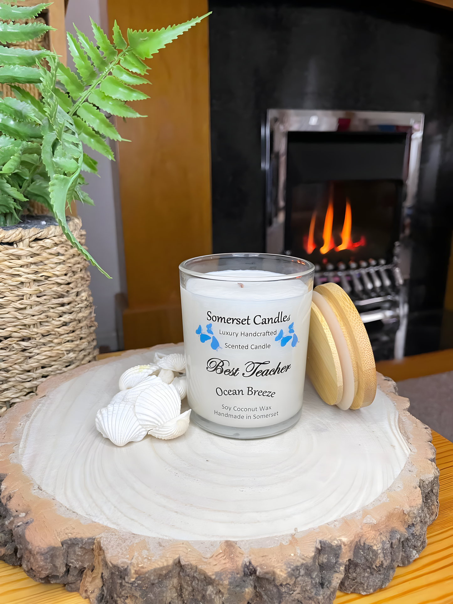 Best Teacher Ocean Breeze scented soy candle displayed in front of a lit fire. The candle is white in colour with bright blue butterflies fluttering above the scent name with a bamboo lid