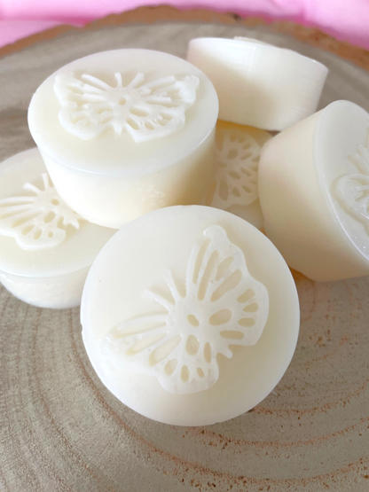 Several round white wax melts with embossed butterfly designs, arranged on a round wooden slice against a pink fabric background. Birthday gift for her.