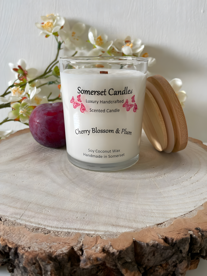 Cherry blossom & plum scented soy candle is displayed on a wooden board with a fresh plum & white cherry blossom. The candle is white in colour with bright red butterflies above the scent name with a lovely bamboo lid lent against the candle