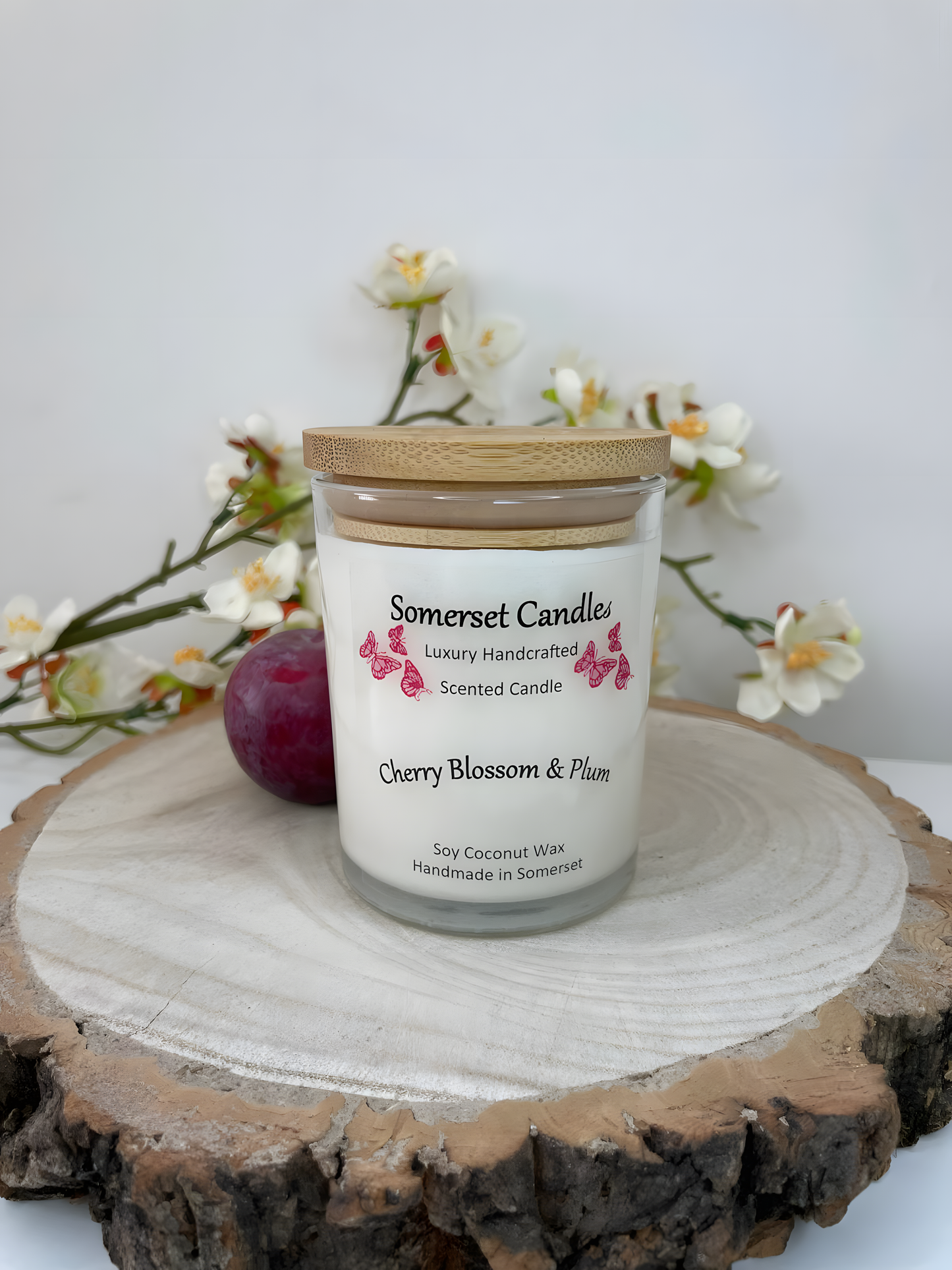 Cherry blossom & plum scented soy candle is displayed on a wooden board with a fresh plum & white cherry blossom. The candle is white in colour with bright red butterflies above the scent name with a lovely bamboo lid.