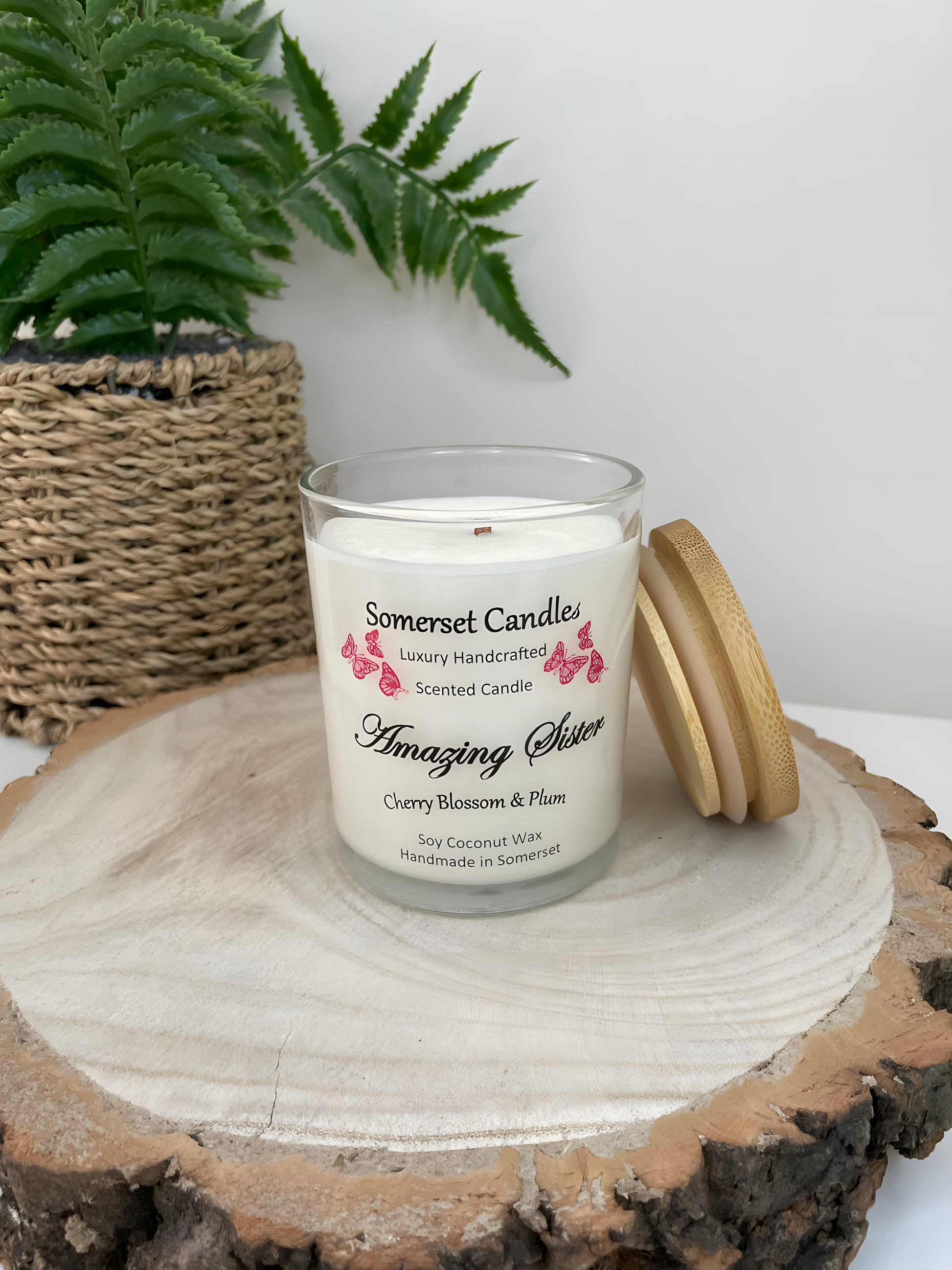 Cherry blossom & plum scented soy candle amazing sister is displayed on a wooden board. The candle is white in colour with bright red butterflies above the scent name with a bamboo lid. The candle jar is clear glass.