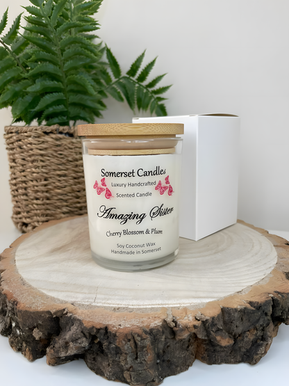 Cherry blossom & plum scented soy candle amazing sister is displayed on a wooden board with a white gift box. The candle is white in colour with bright red butterflies above the scent name with a lovely bamboo lid fitted. Behind there is a plant.