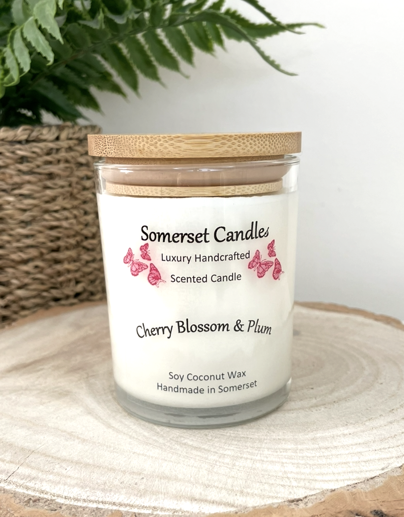 White soy candle in a 7oz clear glass jar with a natural wooden lid. The candle is made from a soy-coconut blend and scented with a delicate mix of cherry blossom and plum. Above the scent name on the label, several red butterflies are displayed, adding a romantic touch. Clean, minimalist design ideal for gifts, home decor or self-care.